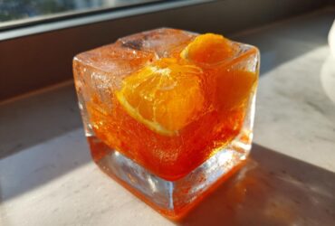 Close-up of an Aperol Spritz popsicle in a square ice cube mold with orange slices inside.