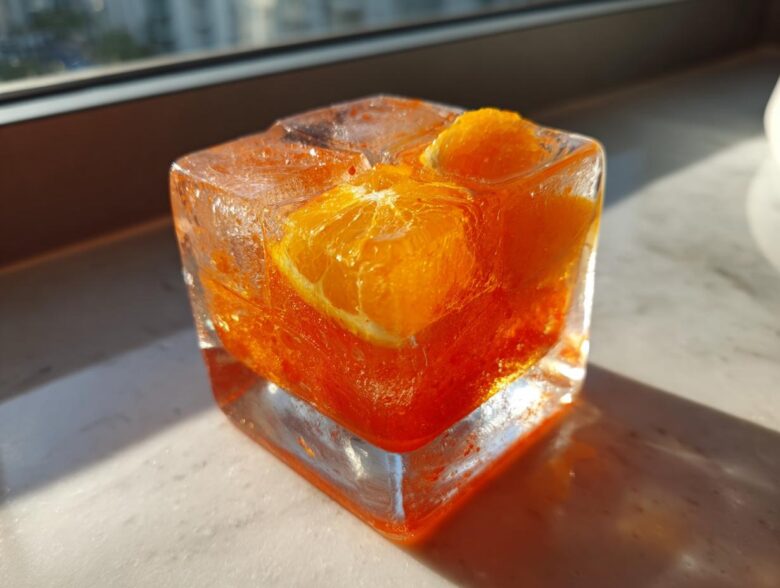 Close-up of an Aperol Spritz popsicle in a square ice cube mold with orange slices inside.