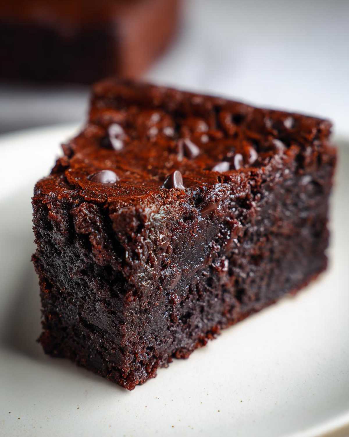A close-up of a rich, fudgy slice of Black Bean Brownies topped with chocolate chips on a white plate.