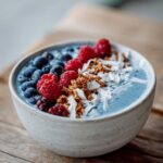 A close-up of a Blue Spirulina Smoothie Bowl topped with fresh blueberries, raspberries, granola, and shredded coconut.
