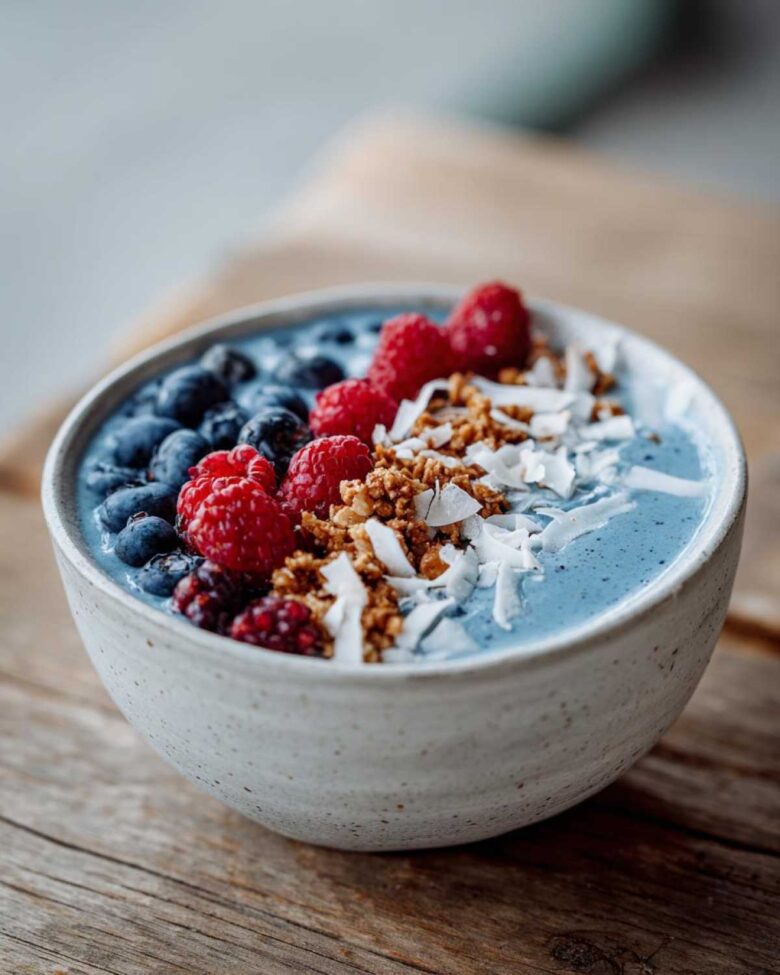 A close-up of a Blue Spirulina Smoothie Bowl topped with fresh blueberries, raspberries, granola, and shredded coconut.