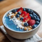 A vibrant Blue Spirulina Smoothie Bowl topped with fresh blueberries, sliced strawberries, granola, and coconut flakes.