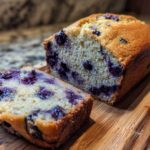 A close-up of a moist Blueberry Banana Bread loaf, with one slice cut and showing plump blueberries.
