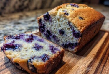 A close-up of a moist Blueberry Banana Bread loaf, with one slice cut and showing plump blueberries.