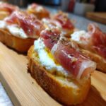 Close-up of delicious Burrata & Prosciutto Crostini appetizer on a wooden board, drizzled with olive oil and herbs.