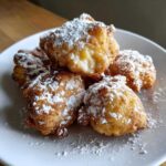 A close-up of golden-brown Cardamom Apple Fritters dusted with powdered sugar on a white plate.