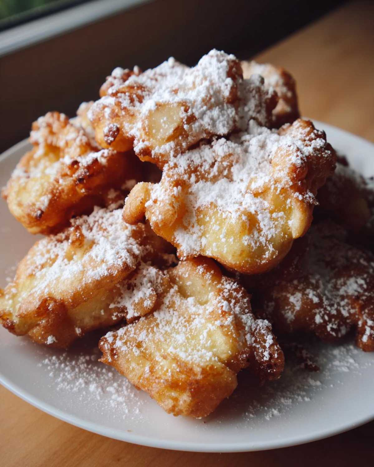 A close-up of a pile of golden-brown Cardamom Apple Fritters generously dusted with powdered sugar on a white plate.