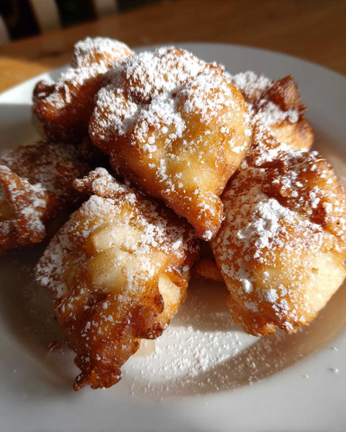 A close-up of golden brown Cardamom Apple Fritters generously dusted with powdered sugar on a white plate.