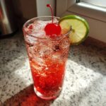 A tall glass filled with a bubbly, red Cherry Limeade Cooler, garnished with a maraschino cherry and a lime slice.