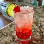 A tall glass filled with a vibrant red Cherry Limeade Cooler, packed with ice, garnished with a maraschino cherry and a lime slice.