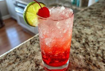 A tall glass filled with a vibrant red Cherry Limeade Cooler, packed with ice, garnished with a maraschino cherry and a lime slice.