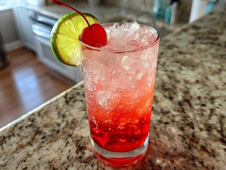 A tall glass filled with a vibrant red Cherry Limeade Cooler, packed with ice, garnished with a maraschino cherry and a lime slice.