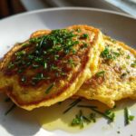Two golden-brown chickpea flour pancakes topped with fresh chives and drizzled with oil on a white plate.