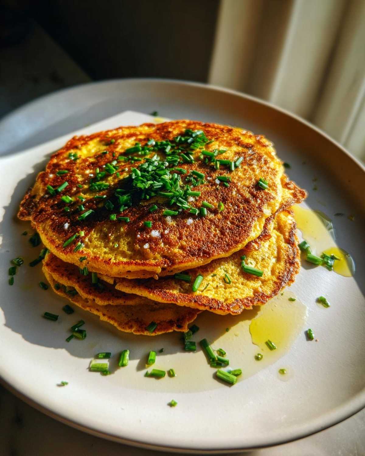 A stack of golden-brown chickpea flour pancakes topped with fresh chives and a drizzle of oil.
