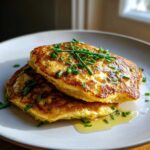 Two golden-brown chickpea flour pancakes stacked on a white plate, drizzled with syrup and topped with fresh chives.