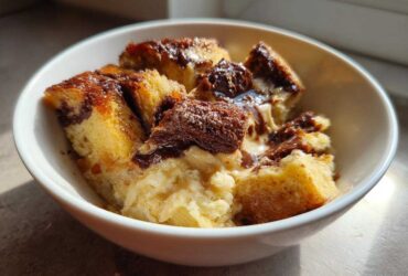 A close-up of a white bowl filled with chunks of Chocolate Babka Bread Pudding, drizzled with chocolate sauce and topped with a dusting of powdered sugar.