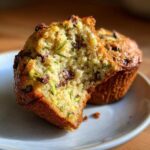 A close-up of a broken chocolate chip zucchini muffin, revealing shredded zucchini and chocolate chips within the moist crumb.