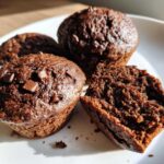 Close-up of moist chocolate protein muffins topped with chocolate chips, one muffin is broken in half to show the texture.