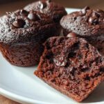Close-up of moist chocolate protein muffins topped with chocolate chips, one muffin is broken in half revealing melted chocolate.