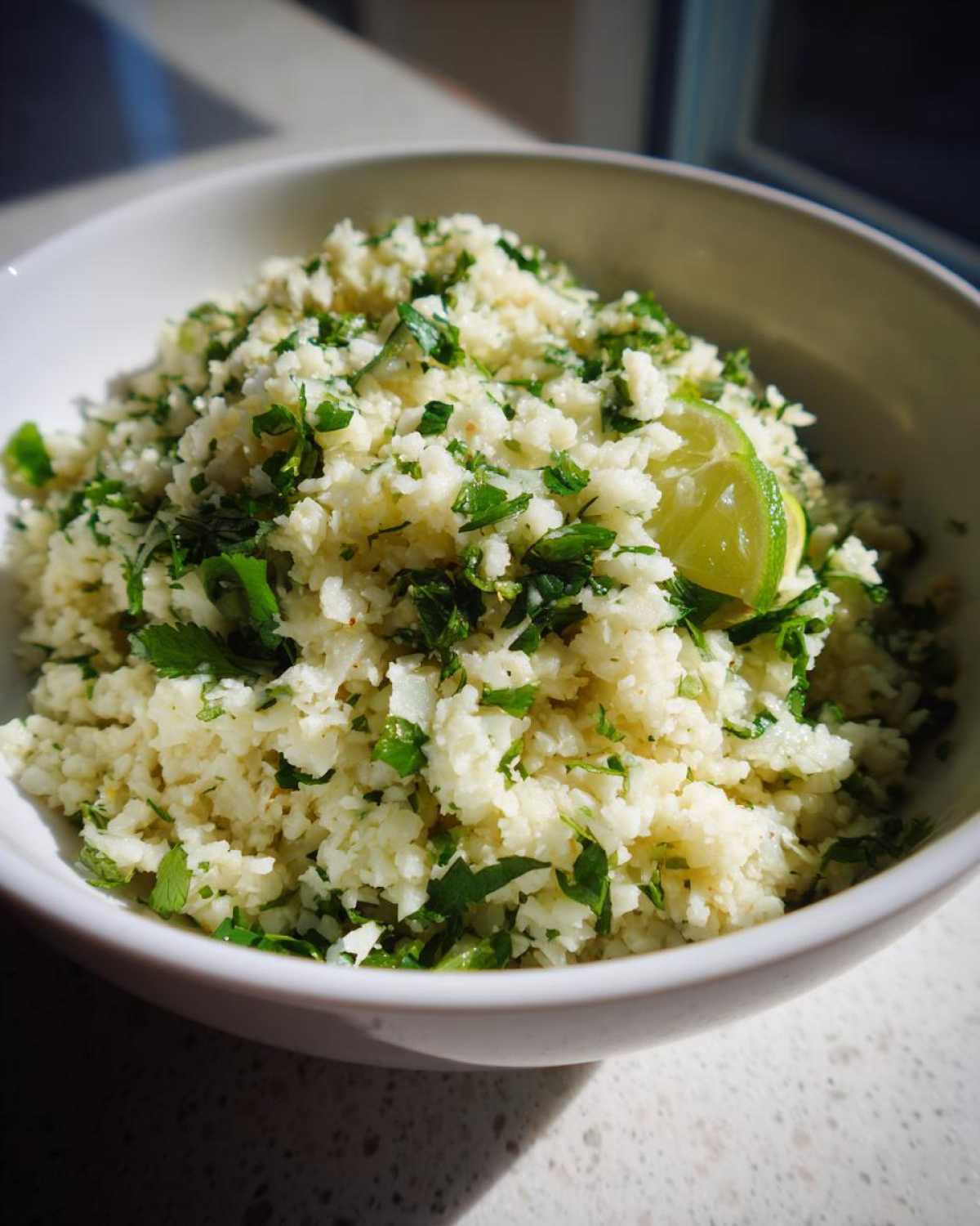 Close-up of a white bowl filled with Cilantro Lime Cauliflower Rice, garnished with fresh cilantro and a lime wedge.