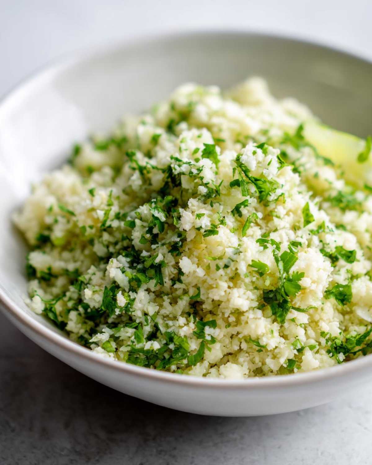 A close-up of a white bowl filled with fluffy Cilantro Lime Cauliflower Rice, garnished with fresh chopped cilantro.