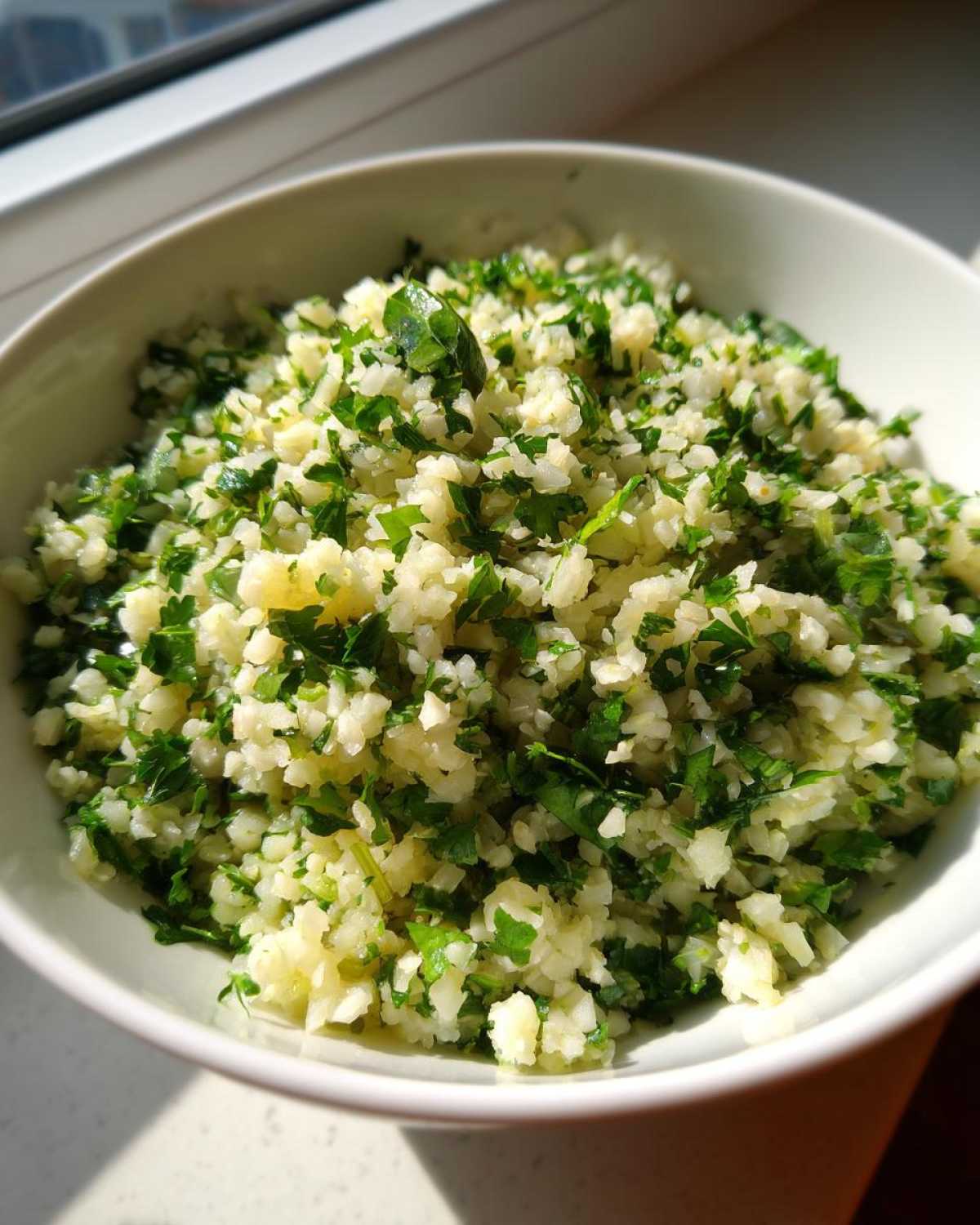 A close-up of a white bowl filled with freshly prepared Cilantro Lime Cauliflower Rice, showcasing the texture and vibrant green herbs.