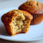 Close-up of a Classic banana muffin with a bite taken out, revealing a moist interior. Another muffin is in the background.