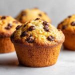 A close-up of a perfectly baked classic chocolate chip muffin, studded with rich chocolate chips.