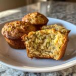 A close-up of moist classic zucchini muffins on a white plate, one muffin is broken in half to show the texture and shredded zucchini inside.