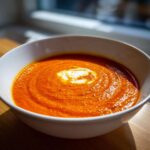 A bowl of vibrant Creamy Roasted Red Pepper Soup with a swirl of cream on top, presented in a white bowl.