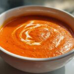 Close-up of a bowl of vibrant Creamy Roasted Red Pepper Soup with a swirl of cream and black pepper.