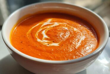 Close-up of a bowl of vibrant Creamy Roasted Red Pepper Soup with a swirl of cream and black pepper.