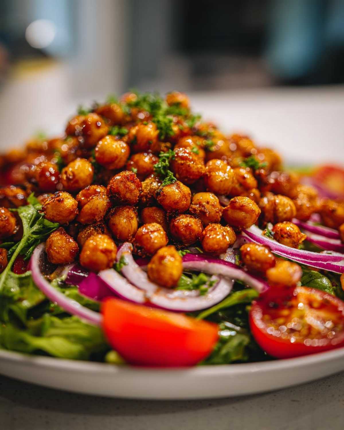 A close-up of a Crispy Chickpea Salad featuring a mound of seasoned chickpeas atop fresh greens, sliced red onion, and cherry tomatoes.