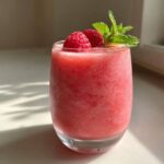 A close-up of a glass filled with pink Frozen Rosé Slushies, garnished with fresh raspberries and mint leaves.