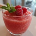 A close-up of a glass filled with a vibrant pink Frozen Rosé Slushie, garnished with fresh raspberries and mint leaves.