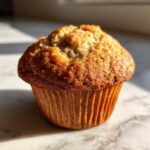 A close-up of a golden-brown gluten-free banana muffin in a fluted paper liner, with visible banana specks.