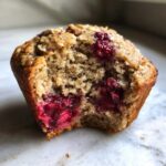 A close-up of a gluten-free protein muffin with a bite taken out, revealing juicy raspberries inside.