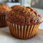 Close-up of a golden-brown gluten-free pumpkin muffin in a paper liner, with more muffins blurred in the background.