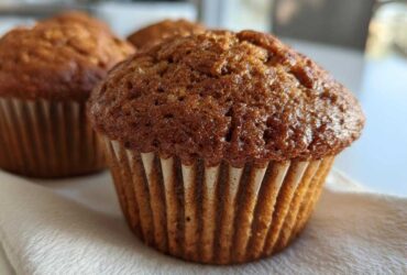 Close-up of a golden-brown gluten-free pumpkin muffin in a paper liner, with more muffins blurred in the background.
