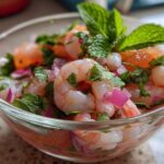 A close-up of Grapefruit Shrimp Ceviche in a clear glass bowl, featuring plump shrimp, red onion, cilantro, and mint.