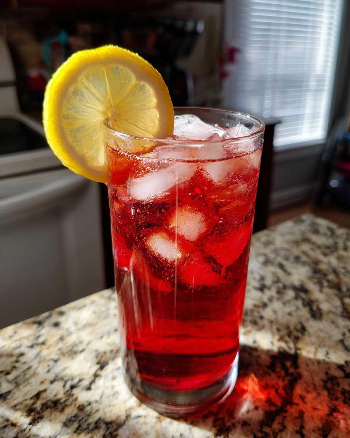 A tall glass filled with vibrant red Hibiscus Iced Tea, ice cubes, and a lemon slice garnish.