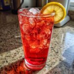 A tall glass filled with vibrant red Hibiscus Iced Tea, ice, and a lemon slice garnish.
