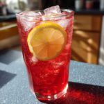 A tall glass filled with vibrant red Hibiscus Iced Tea, packed with ice cubes and garnished with a fresh lemon slice.