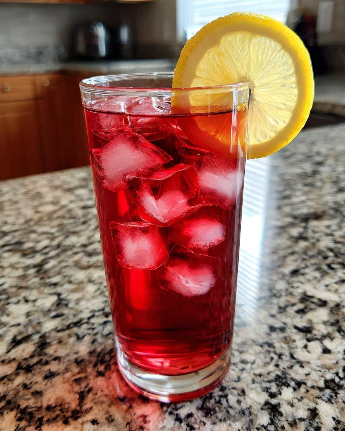 A tall glass filled with vibrant red Hibiscus Iced Tea, packed with ice cubes and garnished with a bright lemon slice.