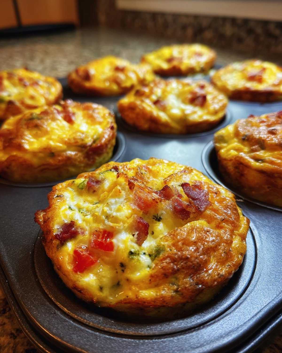 Close-up of freshly baked Keto Egg Muffins in a muffin tin, topped with crumbled bacon and visible pieces of red pepper and broccoli.
