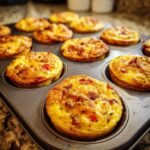 A close-up of freshly baked Keto Egg Muffins in a muffin tin, showing a golden-brown crust with visible pieces of bacon and red bell pepper.