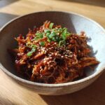 A close-up of delicious vegan Korean BBQ Jackfruit, garnished with sesame seeds and green onions in a rustic bowl.