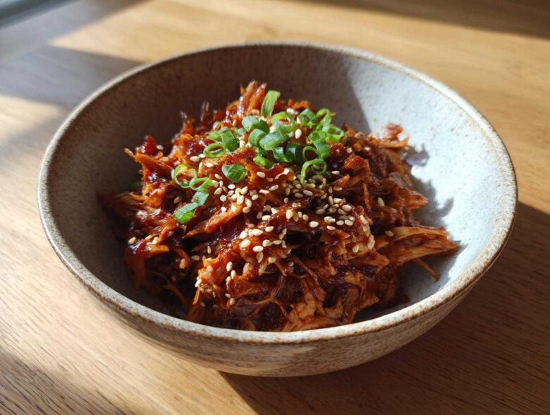 A close-up of delicious vegan Korean BBQ Jackfruit, garnished with sesame seeds and green onions in a rustic bowl.