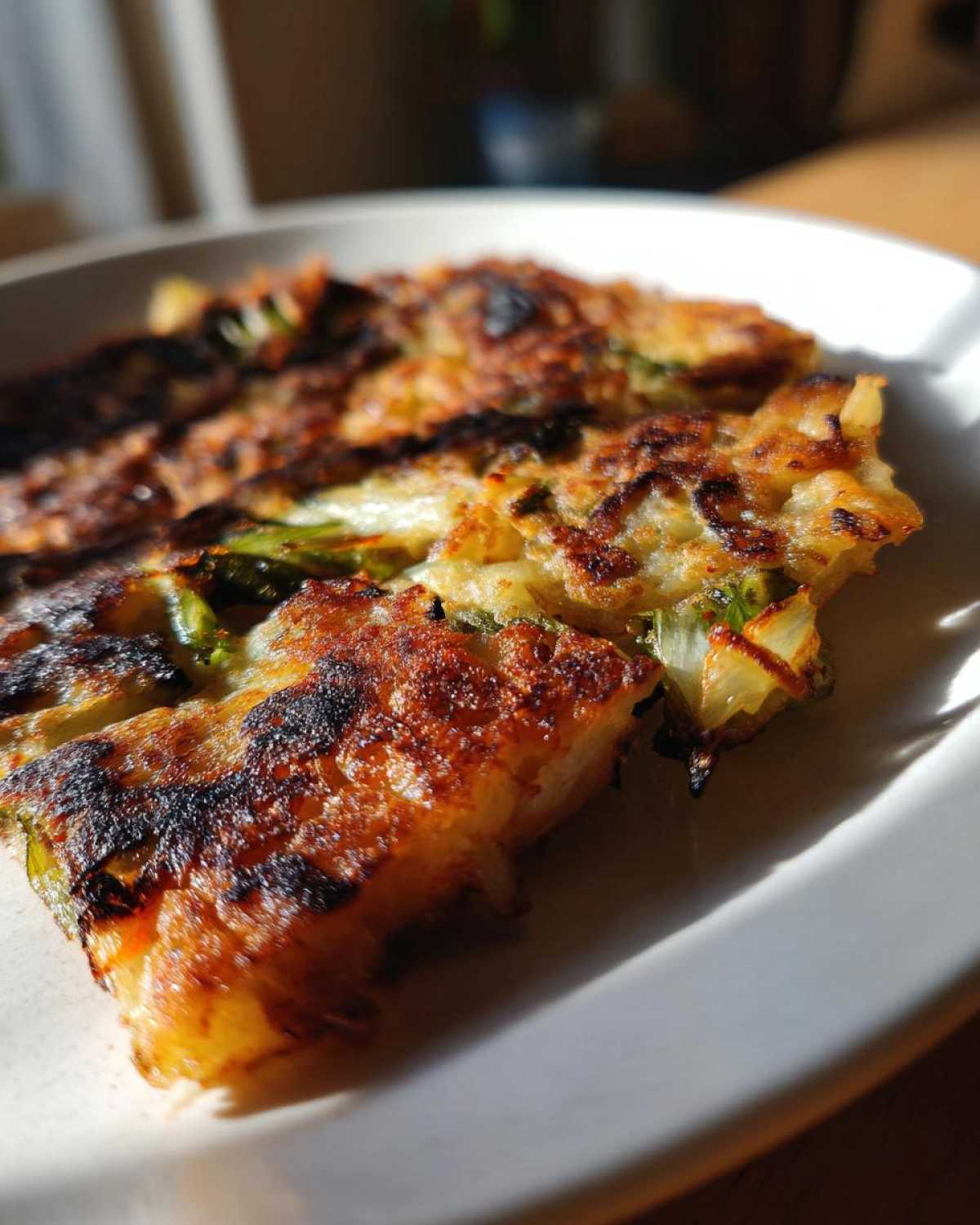 Close-up of a golden-brown, crispy Korean Kimchi Pancake (Pajeon) with visible green onions and a slightly charred edge.