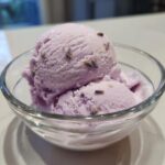 Two scoops of creamy purple Lavender Honey Ice Cream garnished with dried lavender buds in a clear glass bowl.